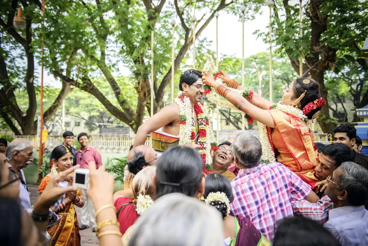Relatives lift a bride and groom for the garland exchange under rain trees, everyone laughing