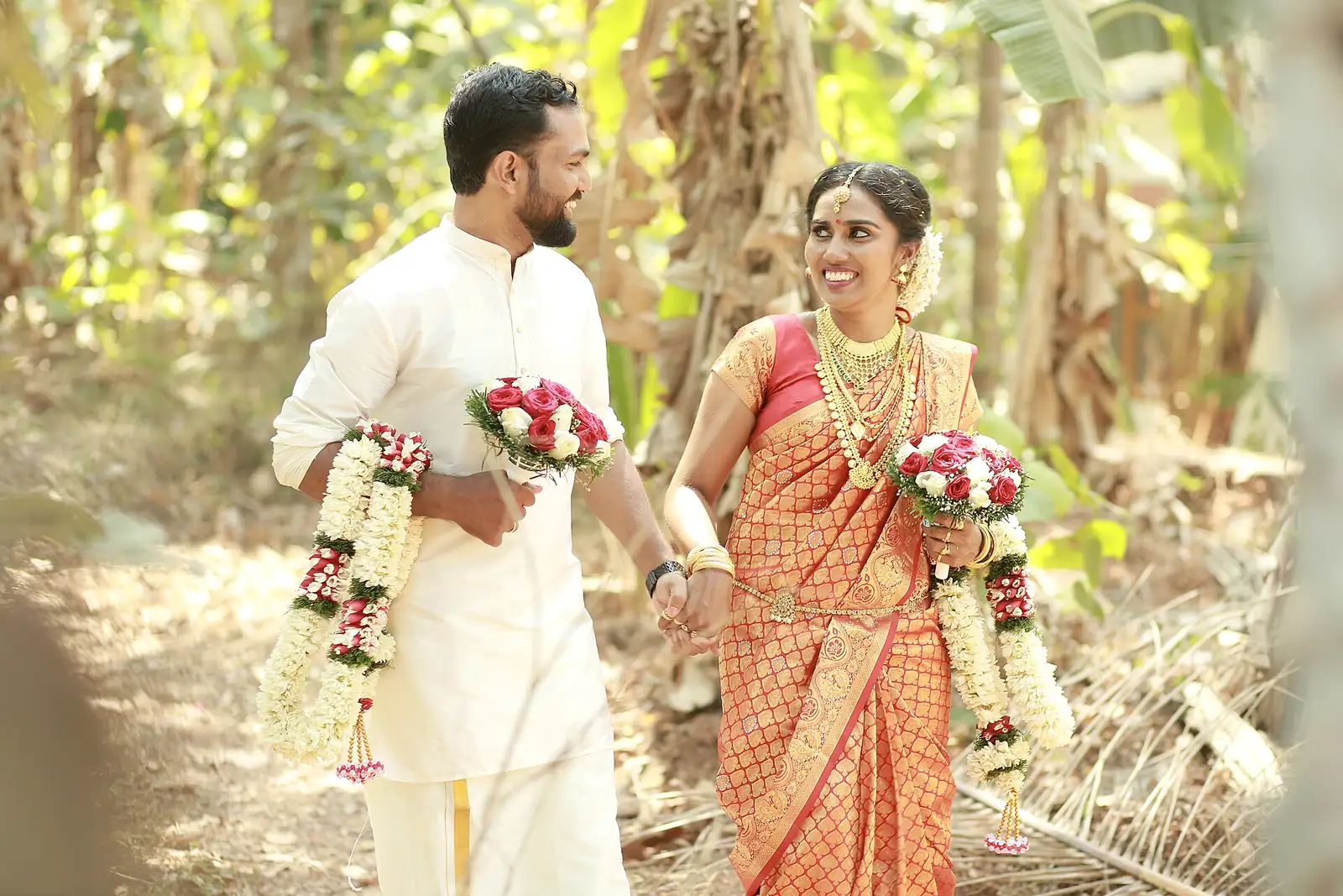 A Kerala bride and groom walk hand in hand through a banana grove, laughing, garlands in hand