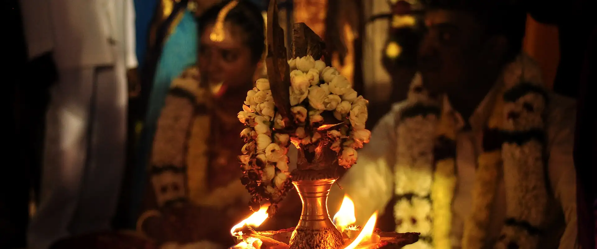A brass kuthu vilakku lit with jasmine at a South Indian wedding, the couple out of focus behind it